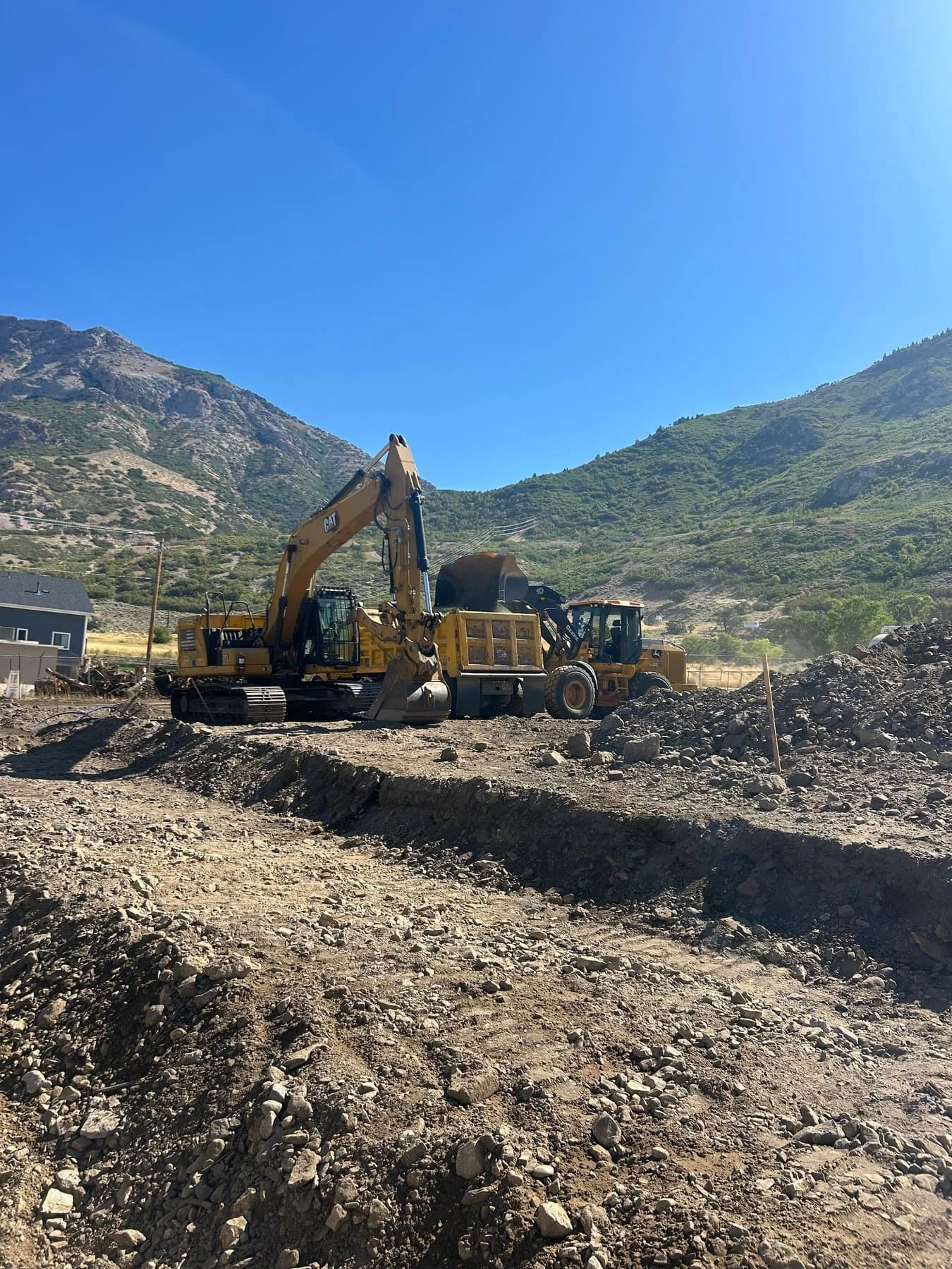 A large CAT excavator and wheel loader excavating a rocky foundation with green mountains behind