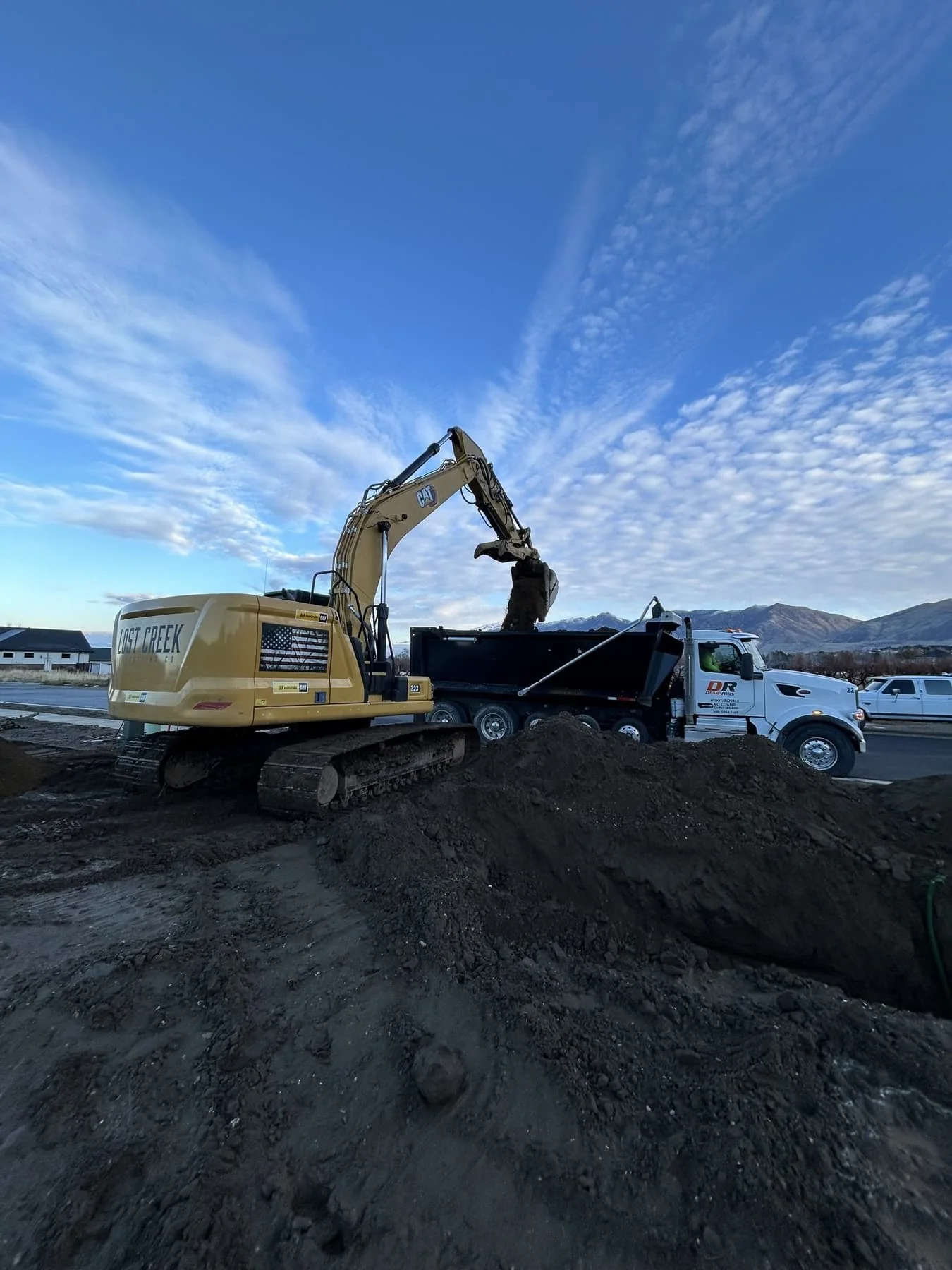 A Lost Creek Contracting CAT excavator loading soil into a dump truck with the Wasatch foothills in the background