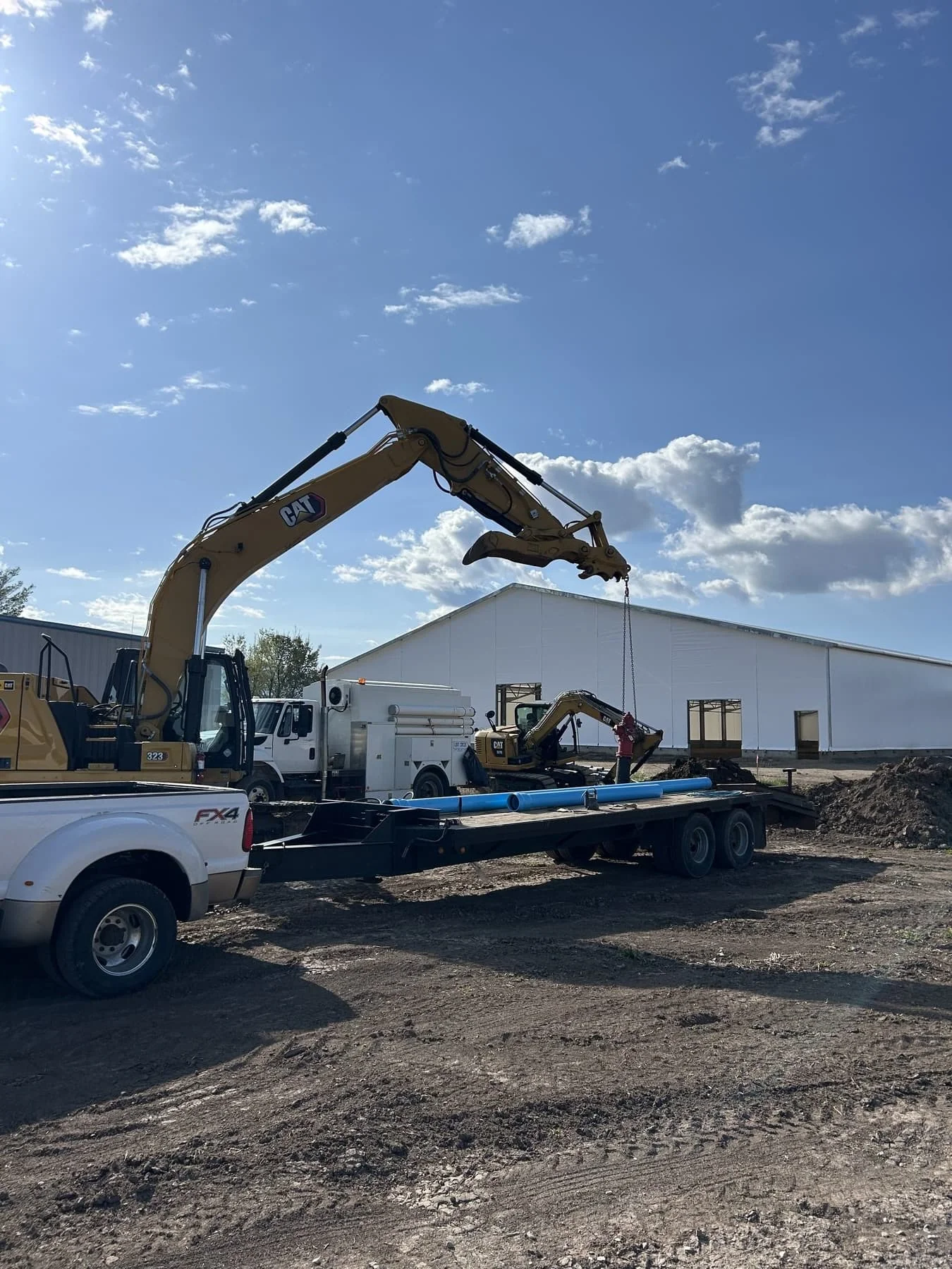 A CAT excavator being loaded onto a flatbed trailer behind a pickup truck at a warehouse site