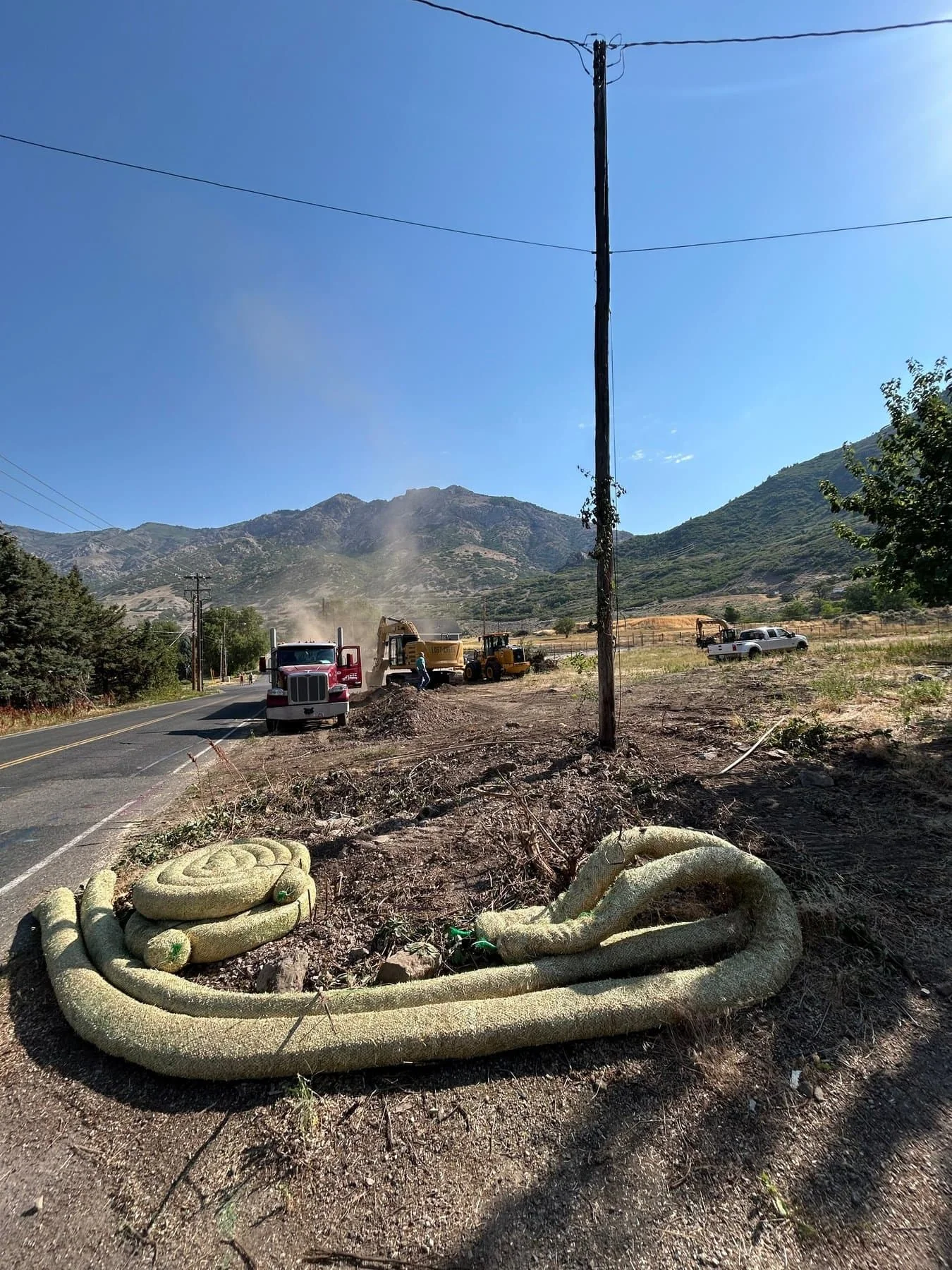 Dump trucks and equipment working a rural roadway with sediment-control wattles laid along the shoulder